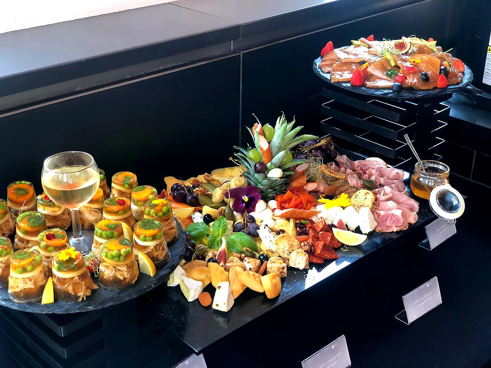 Elegancki stół cateringowy z dekoracyjnymi przekąskami, w tym galaretki w słoiczkach, sery, owoce i wędliny, uzupełniony kieliszkiem białego wina i ananasem.