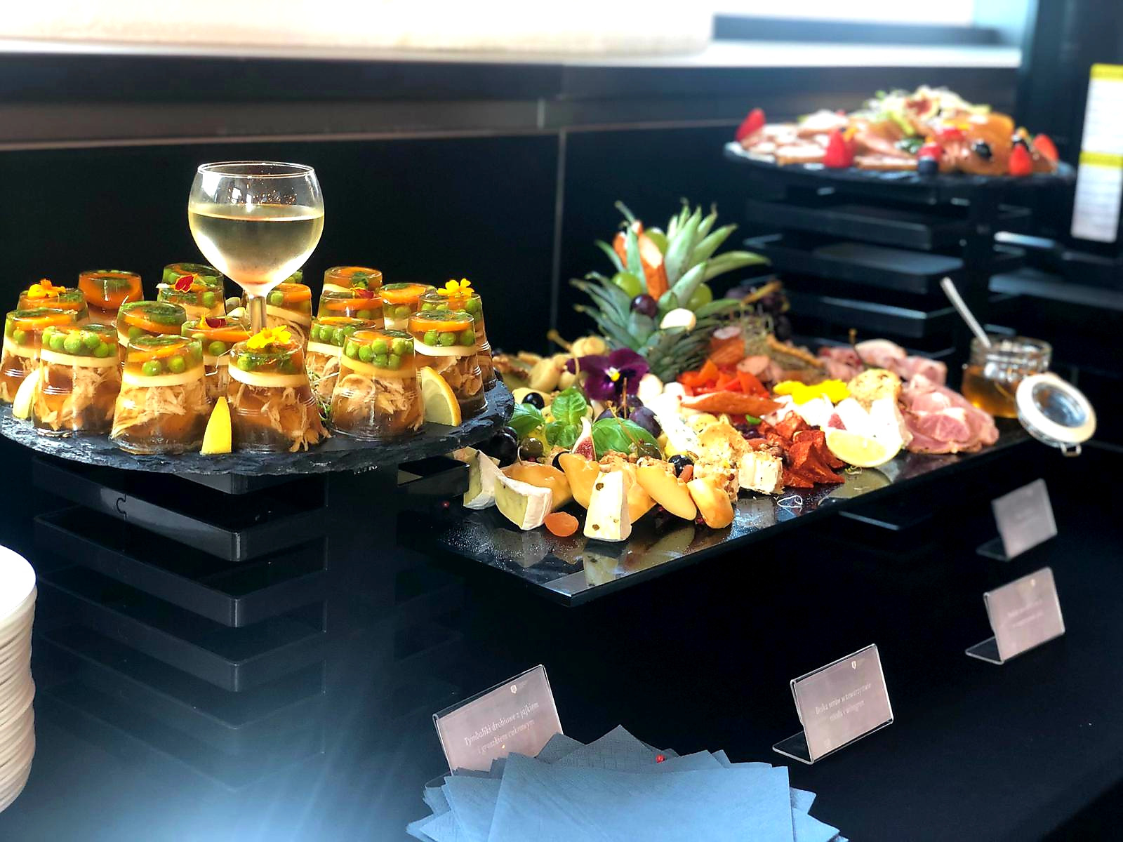 Elegancki stół cateringowy z przekąskami: galaretki drobiowe w słoiczkach, sery, wędliny, owoce i szklanka wina. Dekoracje z kwiatów i owoców. Czarny stół i tło.