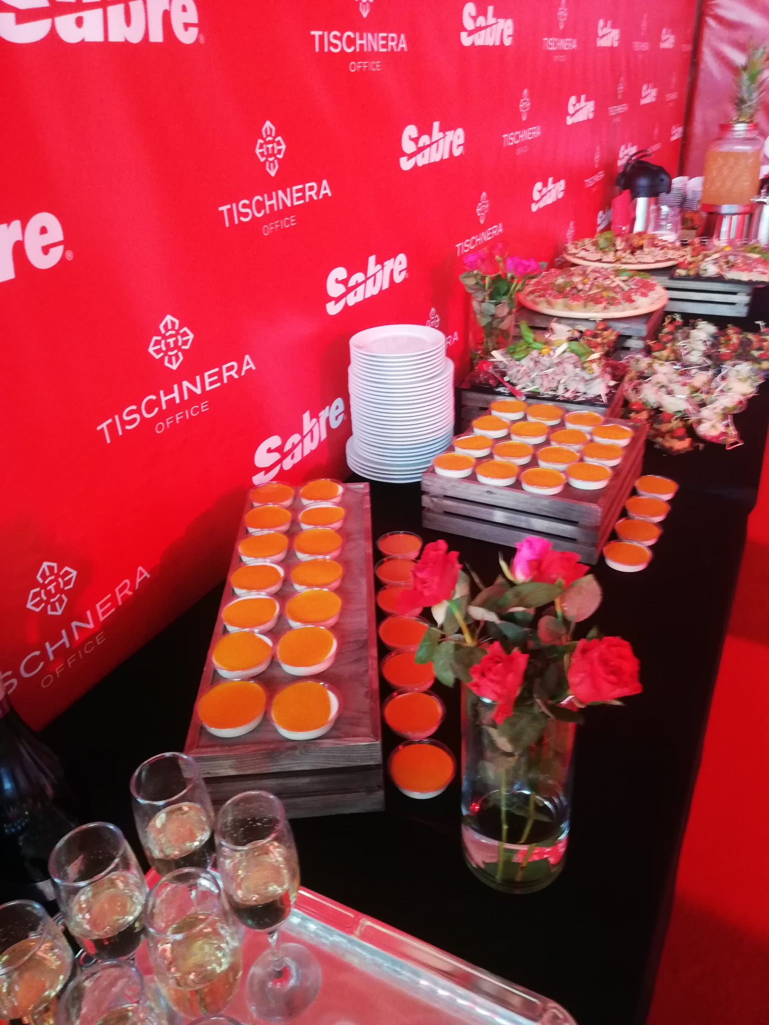 Stół cateringowy z przekąskami, napojami i dekoracjami kwiatowymi na tle czerwonego banera z logo Tischnera Office i Sabre.