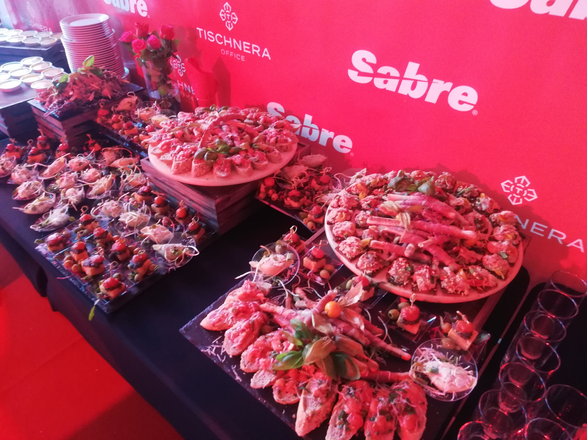 Elegancki stół cateringowy z różnorodnymi przekąskami, w tym bruschetta i mini-kanapeczki, na tle czerwonej ściany z logo Tischnera Office i Sabre.