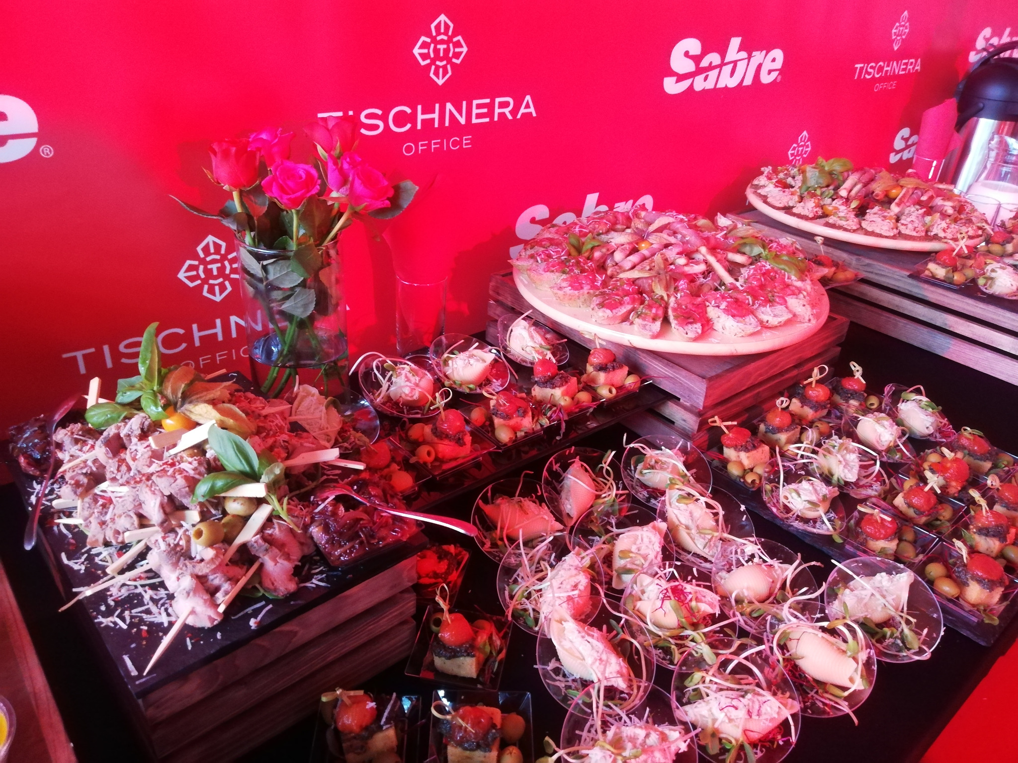 Bogato zastawiony stół z przekąskami na imprezie firmowej w Krakowie. Różnorodne finger food, kompozycje mięsne, dekoracje kwiatowe i logo firmowe w tle.