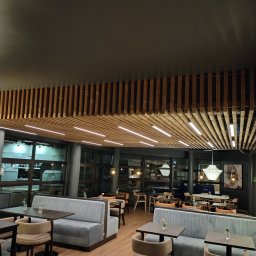 Rafał Stypułkowski - Nowoczesne wnętrze kawiarni z drewnianą konstrukcją sufitową, szarymi sofami i drewnianą podłogą. Widoczne stoliki z kwiatami i designerskie lampy.