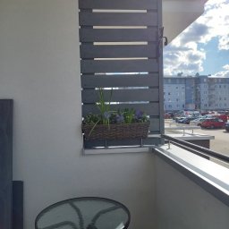 Rafał Stypułkowski - Szara, drewniana zabudowa balkonu z poziomych desek, donica z kwiatami, stolik i krzesło na balkonie w bloku, słoneczny dzień.