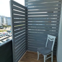 Rafał Stypułkowski - Szara, drewniana zabudowa balkonu z poziomych desek. Krzesło w rogu. Widok na osiedle zza przesłony. Parkiet na podłodze.