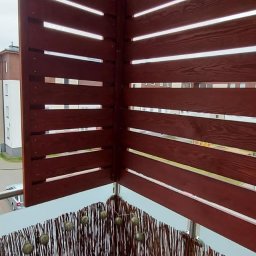 Rafał Stypułkowski - Zabudowa balkonu z brązowych, drewnianych paneli z poziomymi szczelinami i wiklinową osłoną. Widok na fragment budynku w tle.