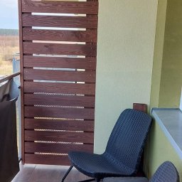 Rafał Stypułkowski - Drewniana zabudowa balkonu z poziomych desek w kolorze ciemnego brązu, z widocznymi dwoma krzesłami z tworzywa sztucznego na drewnianej podłodze.