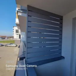 Nowoczesna zabudowa balkonu z szarych, poziomych paneli, widok z zewnątrz na tle błękitnego nieba i fragmentu osiedla. Minimalistyczny design.