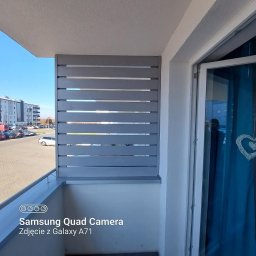 Rafał Stypułkowski - Szara, drewniana zabudowa balkonu z poziomych desek, widok z wewnątrz balkonu, na zewnątrz budynki mieszkalne i parking. Częściowo otwarte drzwi balkonowe.