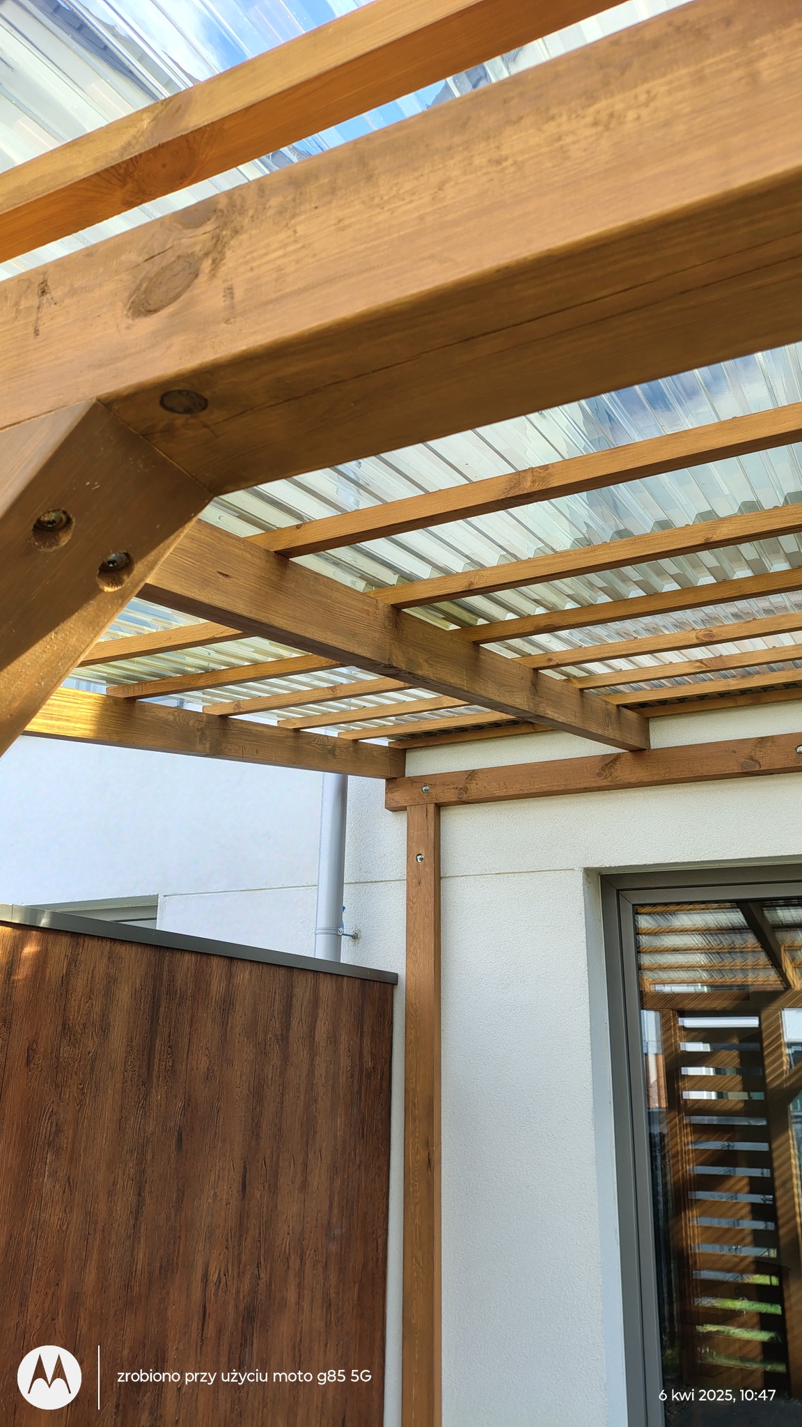 Drewniana pergola z przezroczystym zadaszeniem, widok od dołu, ukazujący strukturę i połączenia belek. Fragment drewnianego ogrodzenia w tle.