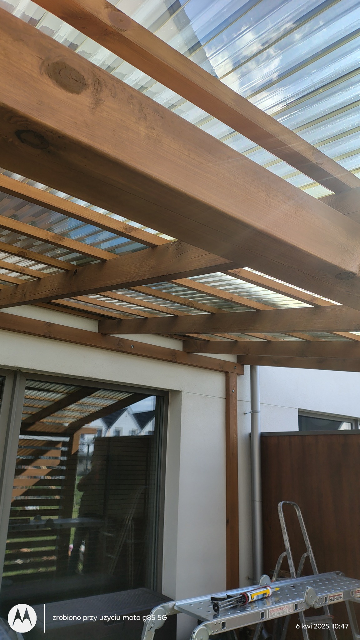 Drewniana konstrukcja z przezroczystym zadaszeniem, widok z dołu. Aluminiowa drabina z narzędziami obok ściany budynku. W tle widoczne okno.