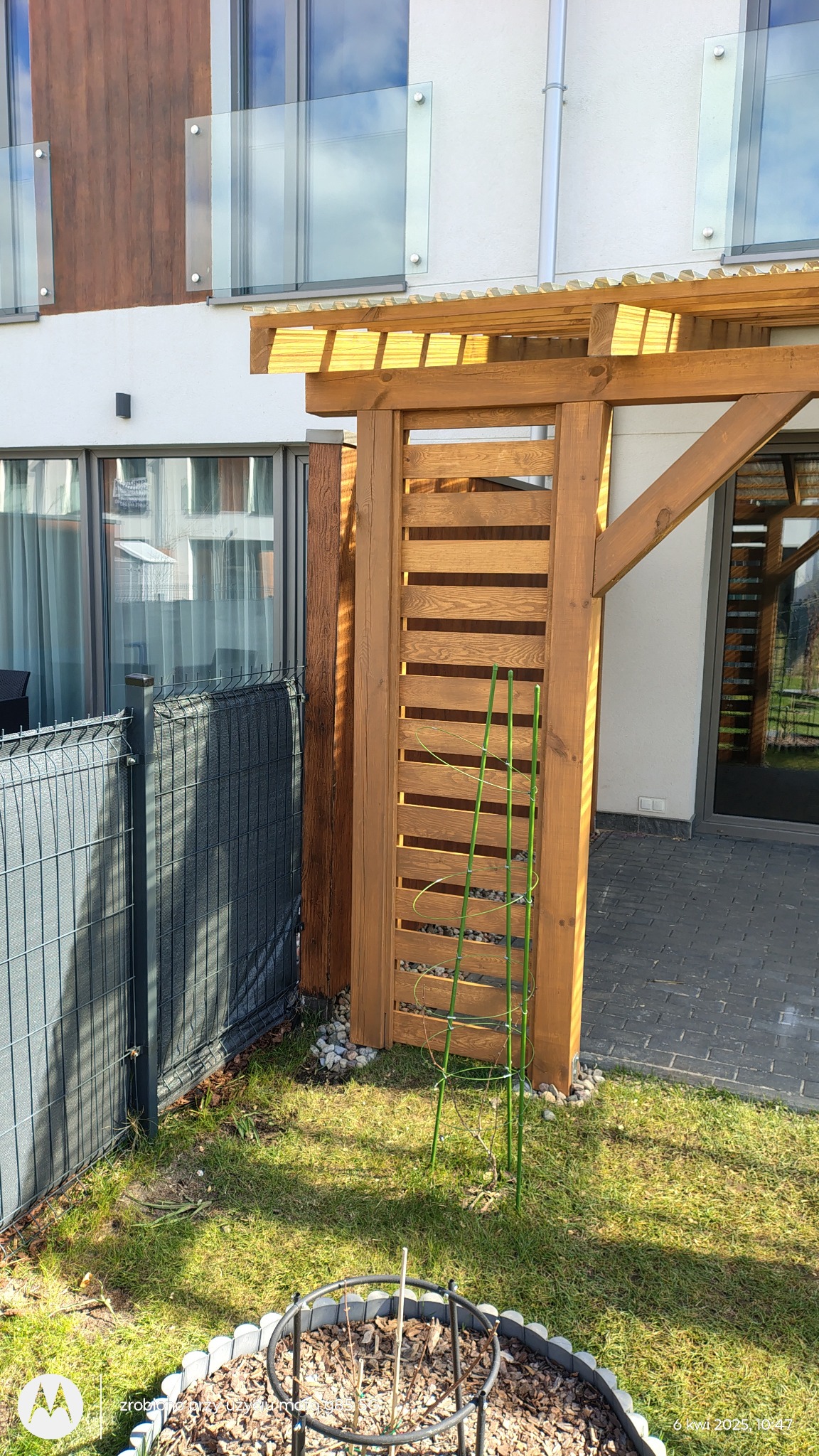 Drewniana pergola z poziomymi listwami, przylegająca do budynku z przeszklonym balkonem, obok siatka ogrodzeniowa i trawnik z młodymi roślinami.