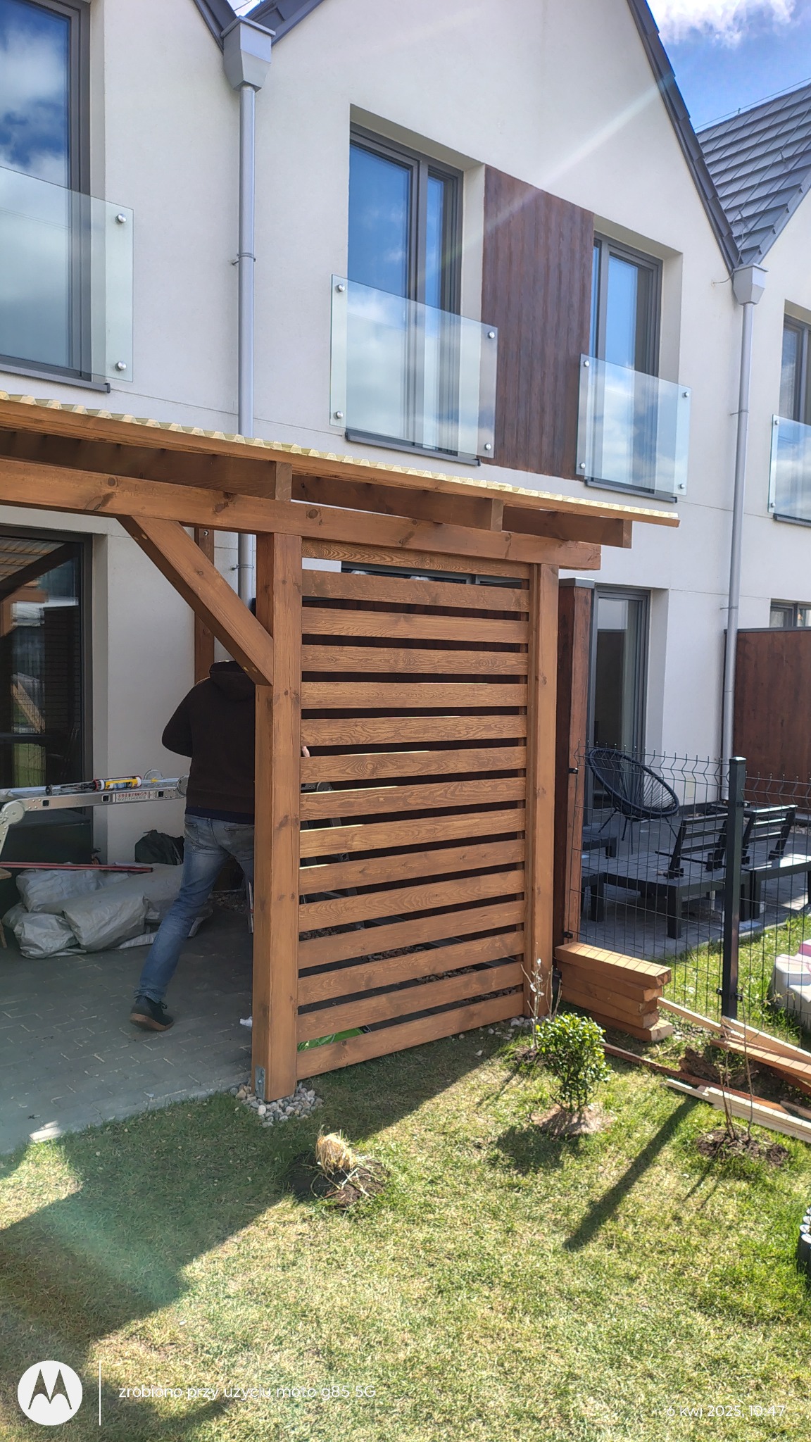 Montaż drewnianej pergoli z poziomymi listwami przy nowoczesnym domu. Widoczny pracownik w trakcie instalacji. Trawa i kostka brukowa w otoczeniu.