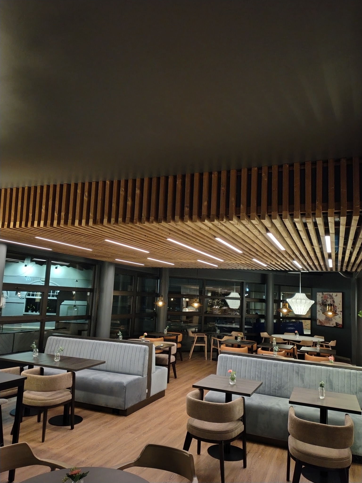 Nowoczesne wnętrze kawiarni z drewnianą konstrukcją sufitową, szarymi sofami i drewnianą podłogą. Widoczne stoliki z kwiatami i designerskie lampy.