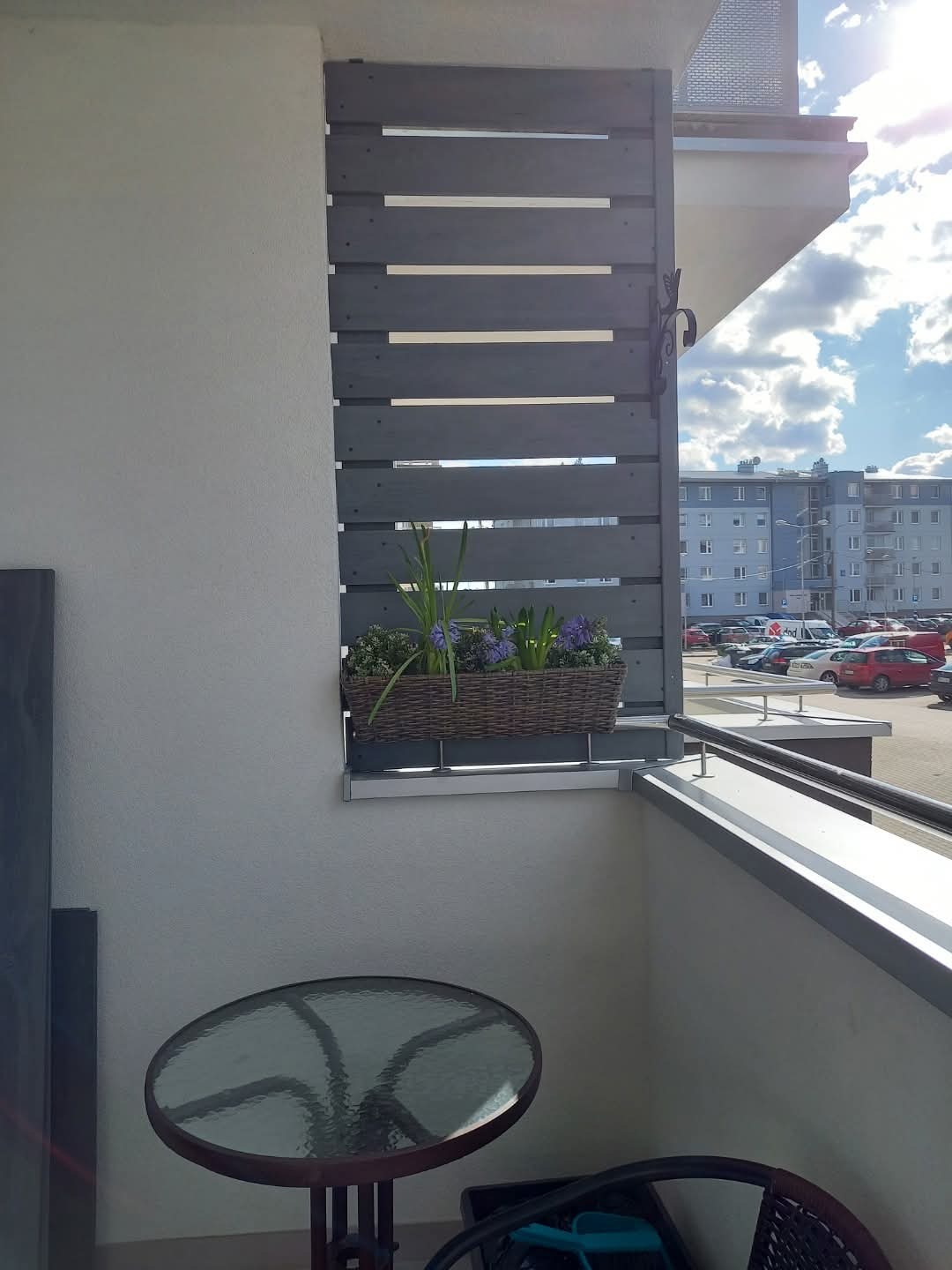 Szara, drewniana zabudowa balkonu z poziomych desek, donica z kwiatami, stolik i krzesło na balkonie w bloku, słoneczny dzień.