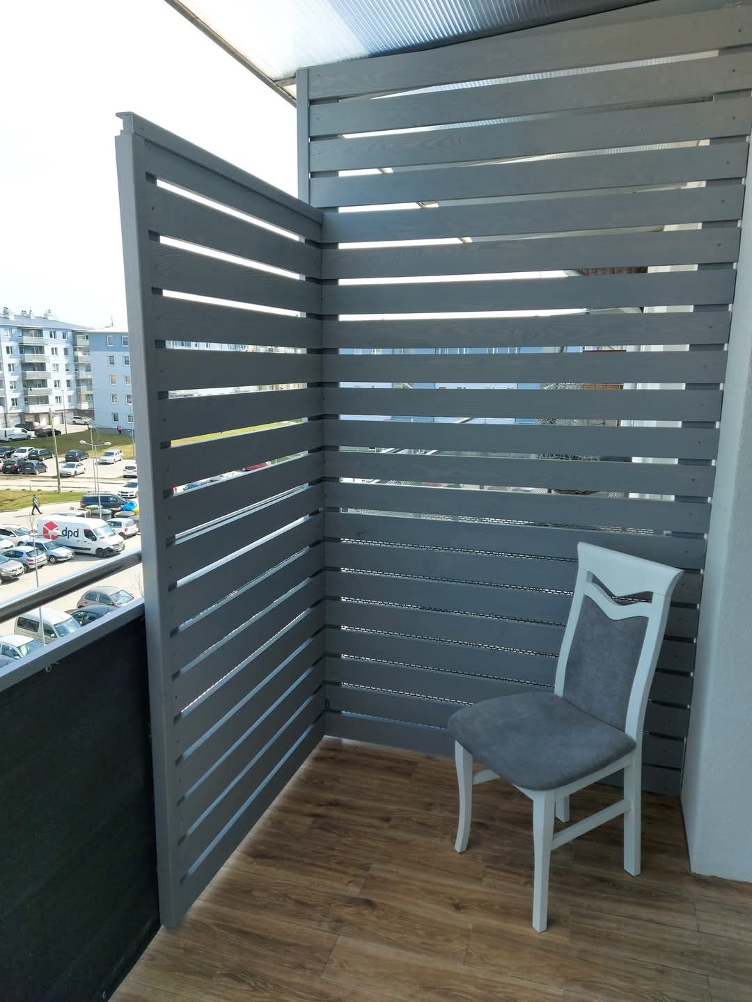 Szara, drewniana zabudowa balkonu z poziomych desek. Krzesło w rogu. Widok na osiedle zza przesłony. Parkiet na podłodze.