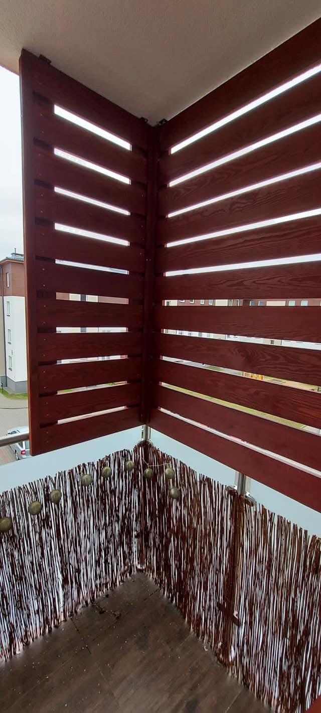 Zabudowa balkonu z brązowych, drewnianych paneli z poziomymi szczelinami i wiklinową osłoną. Widok na fragment budynku w tle.