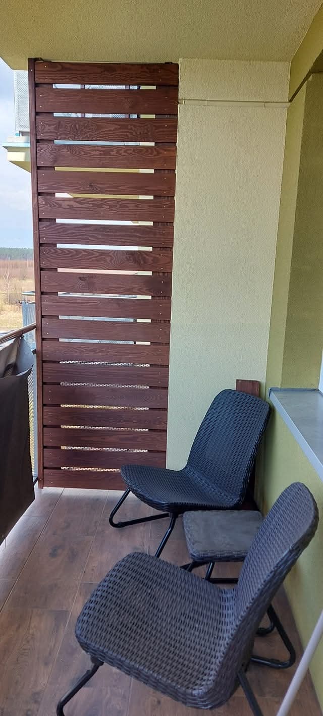 Drewniana zabudowa balkonu z poziomych desek w kolorze ciemnego brązu, z widocznymi dwoma krzesłami z tworzywa sztucznego na drewnianej podłodze.