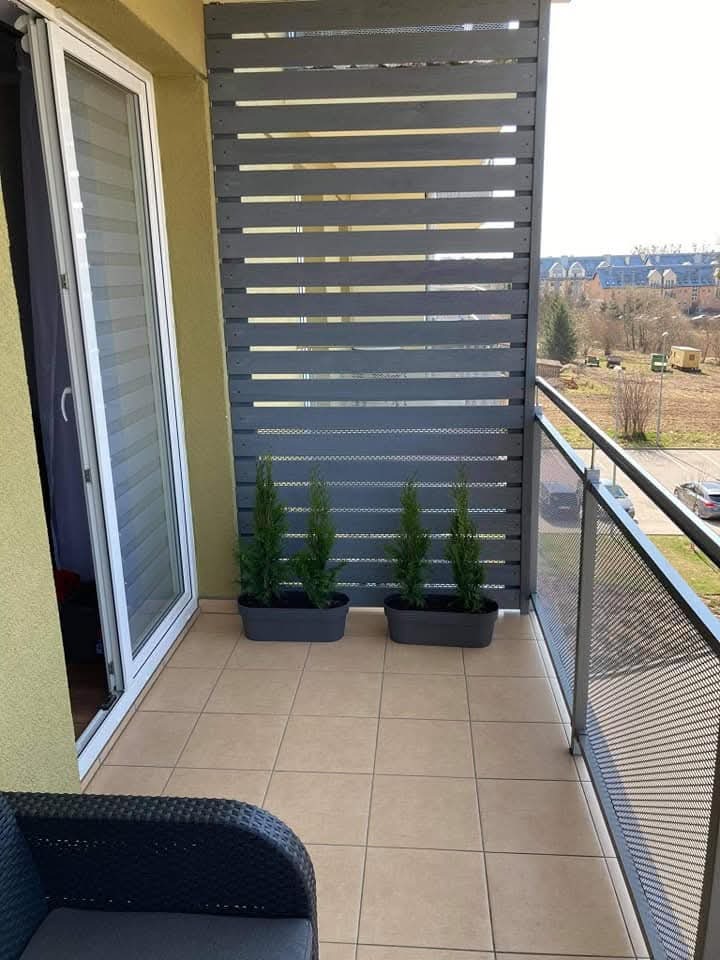 Nowoczesna zabudowa balkonu z szarych, poziomych desek. Na podłodze beżowe płytki, a w tle widok na budynki i drzewa. Ustawione donice z zielonymi roślinami.