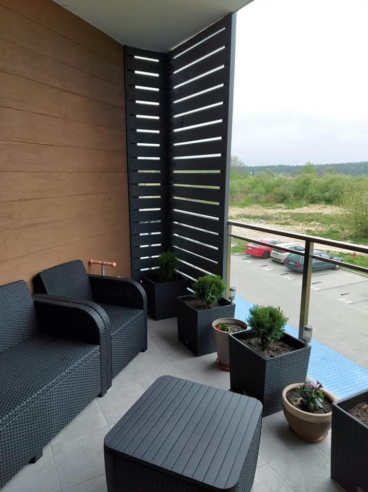 Nowoczesna zabudowa balkonu z elementami drewnopodobnymi i ażurową ścianką. Komplet mebli ogrodowych w kolorze czarnym, donice z roślinami. Widok na parking.