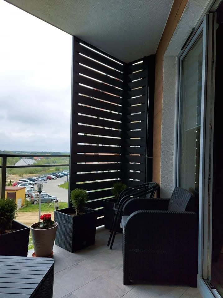 Zabudowa balkonu z ciemnego drewna z poziomymi szczeblami, czarne rattanowe meble, donice z roślinami, widok na parking i zieleń w tle.