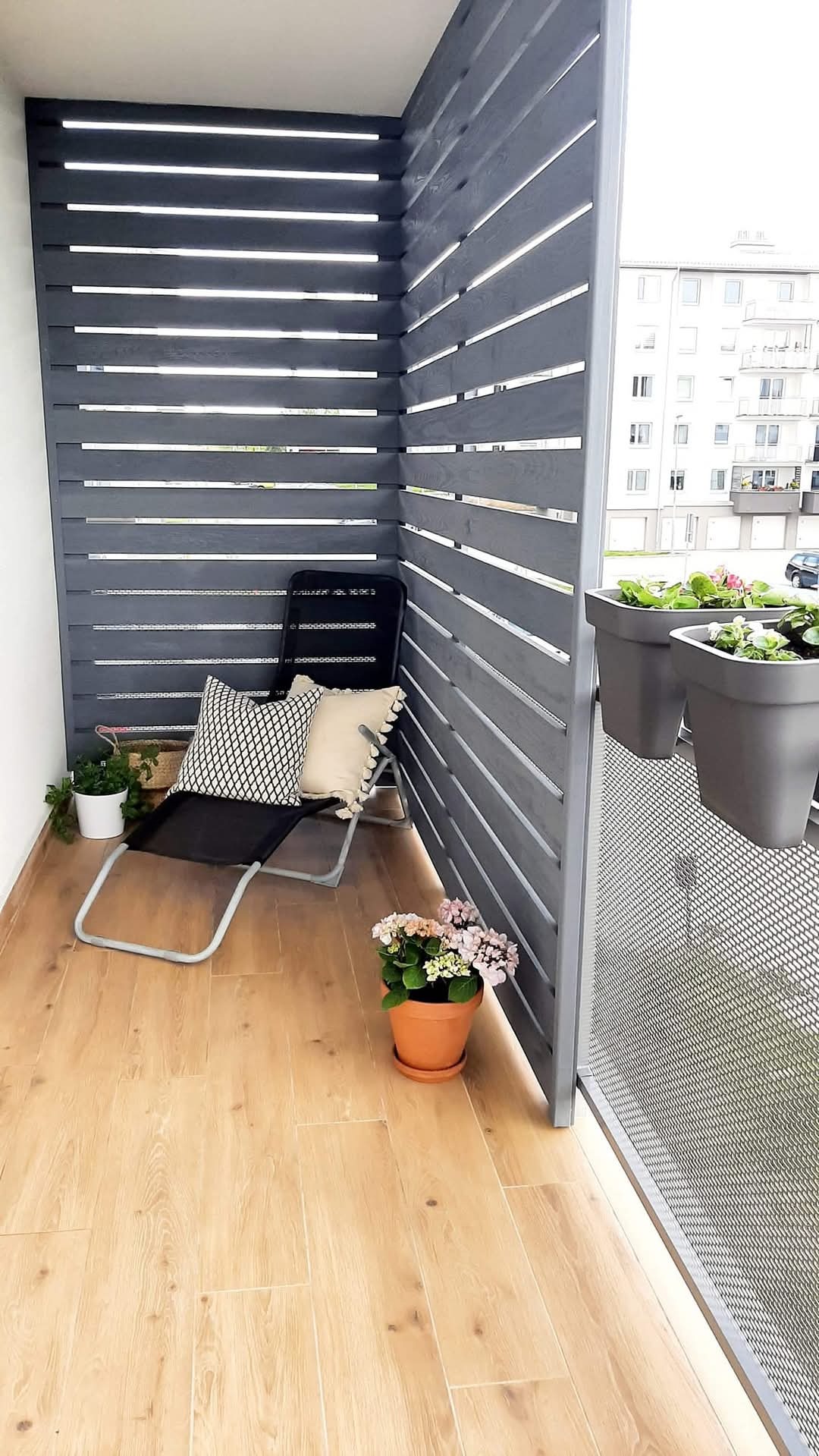 Nowoczesna zabudowa balkonu z szarych, drewnianych paneli, krzesło i kwiaty. Jasna podłoga imitująca drewno. Balkon w bloku.