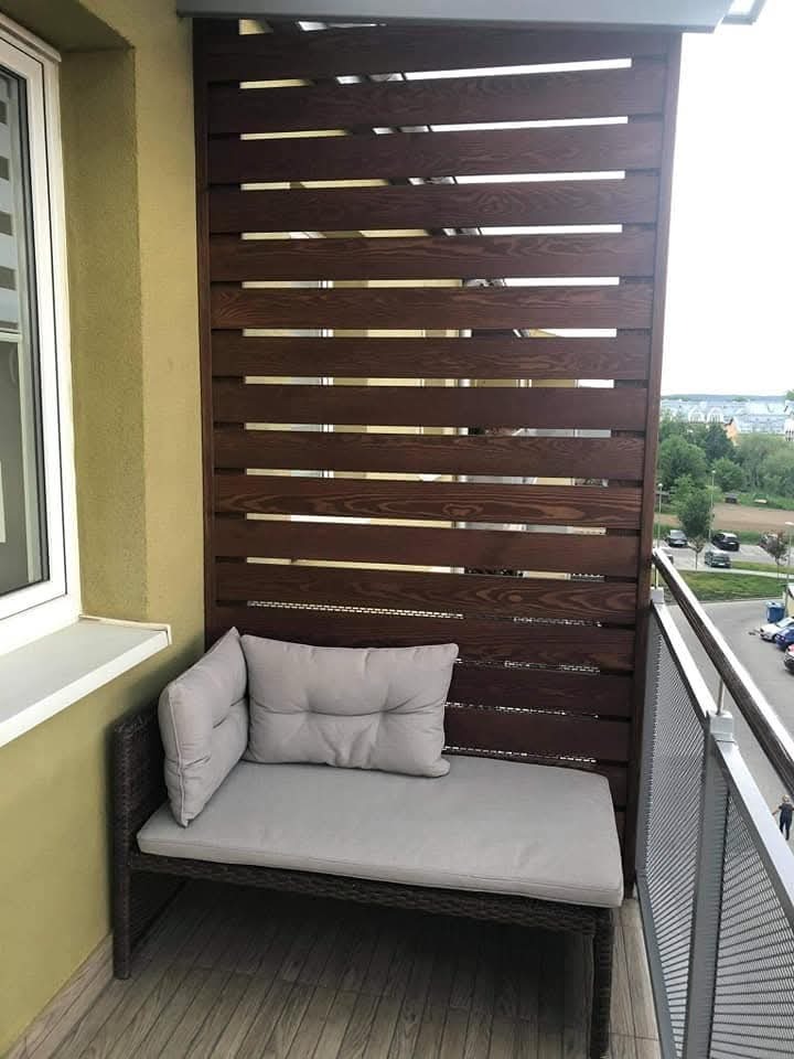 Zabudowa balkonu z drewnianymi panelami w odcieniach brązu, szara sofa z poduszkami, widok na miasto w tle i metalowa balustrada balkonu.