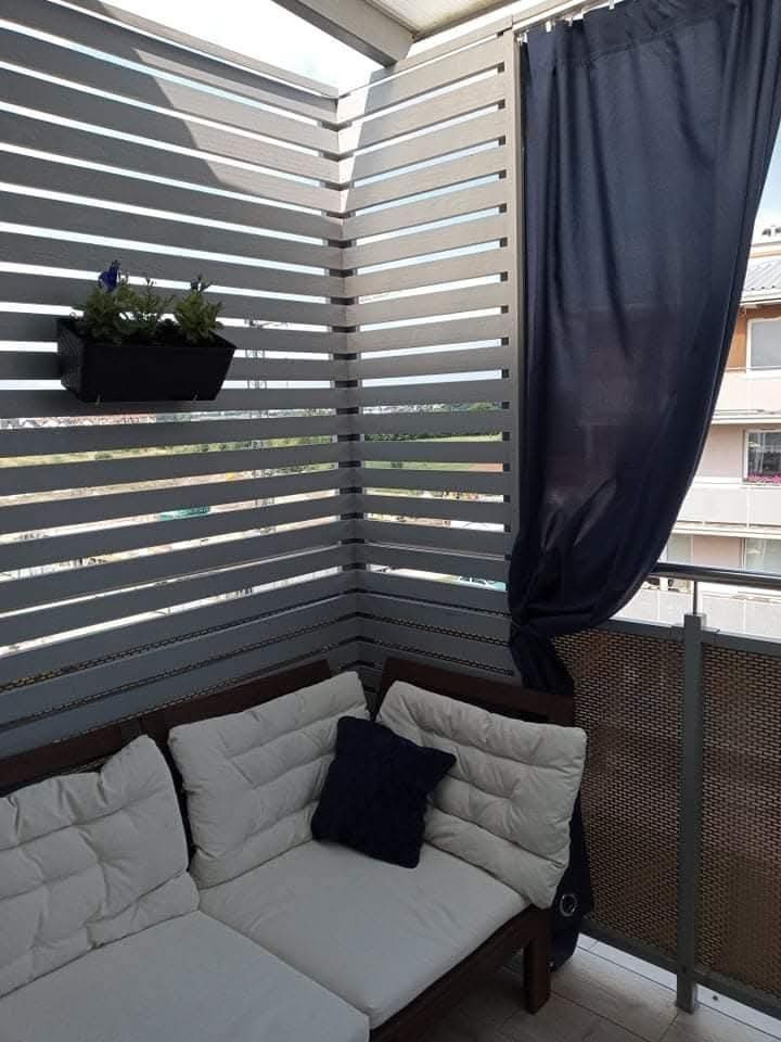 Zabudowany balkon z drewnianą konstrukcją, zasłoną i kwiatami w doniczce. Widoczny narożny zestaw wypoczynkowy z poduszkami. Jasne, naturalne światło.