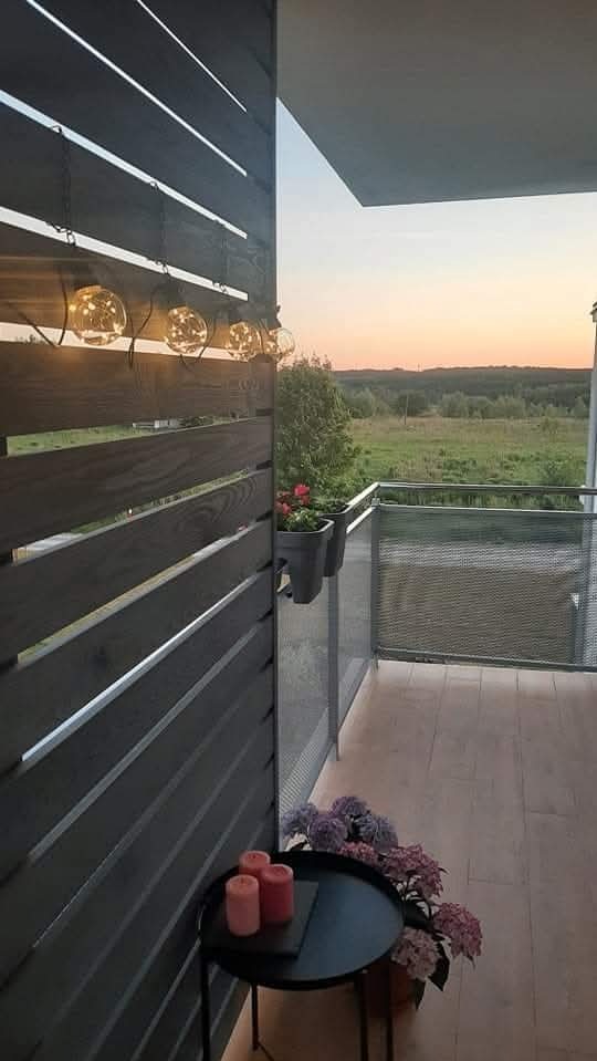 Osłonięty balkon z drewnianą ścianką, ozdobiony lampkami i kwiatami, z widokiem na zielony krajobraz o zachodzie słońca. Przytulna aranżacja z elementami dekoracyjnymi.