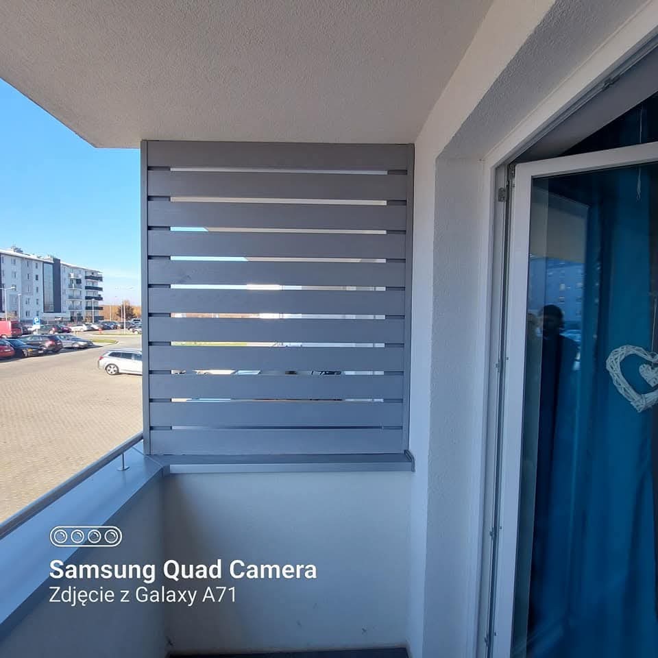 Szara, drewniana zabudowa balkonu z poziomych desek, widok z wewnątrz balkonu, na zewnątrz budynki mieszkalne i parking. Częściowo otwarte drzwi balkonowe.