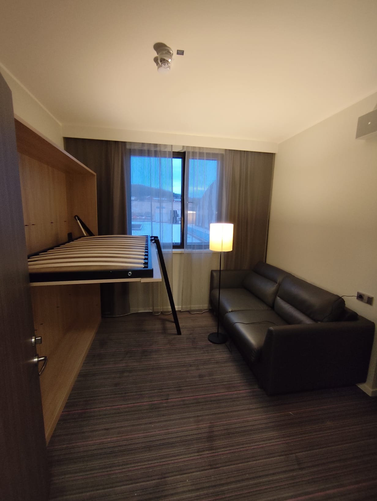 Wnętrze pokoju z rozkładaną szafą-łóżkiem, czarną sofą i lampą podłogową. Widok na okno z szarymi zasłonami. Ciemna wykładzina podłogowa w paski.