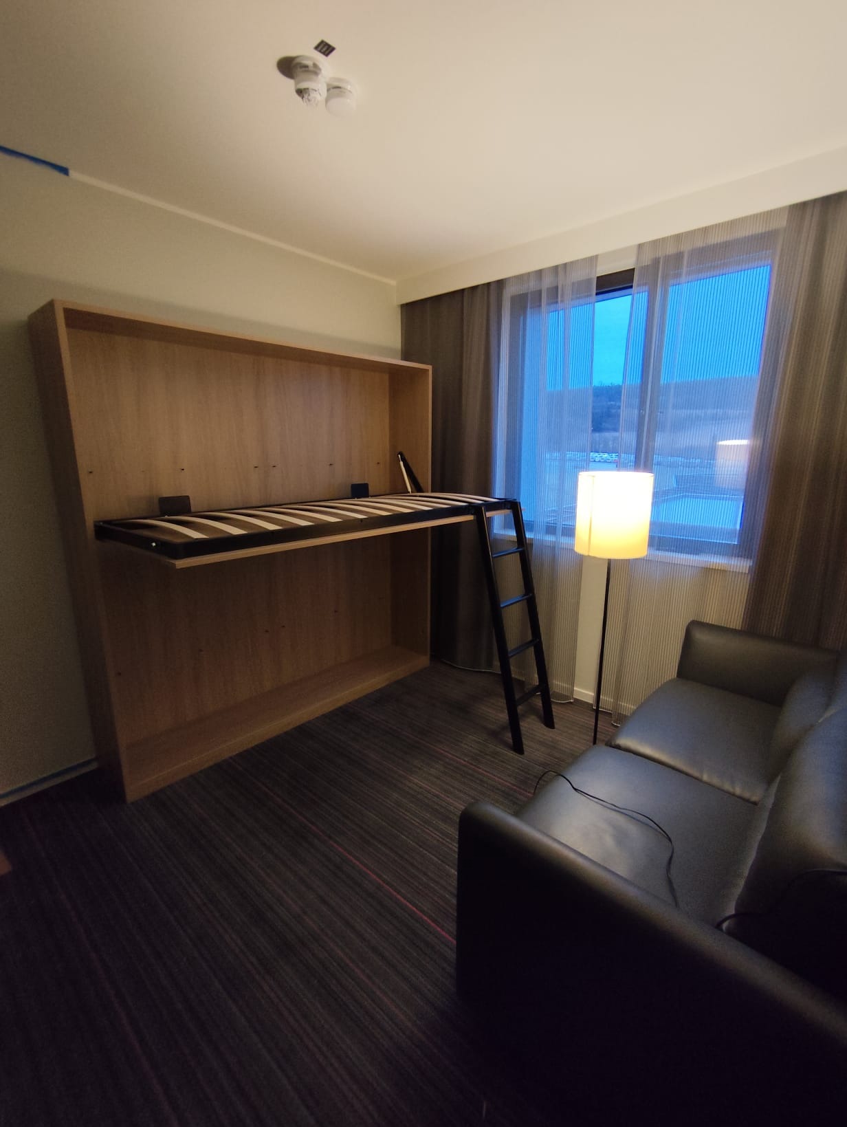 Rozkładane łóżko w zabudowie z drabinką w pokoju hotelowym, obok sofa i lampa podłogowa. Widok na okno z zasłonami.