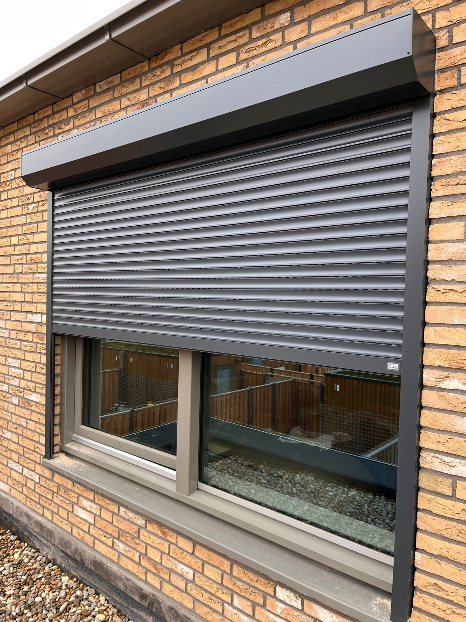Szara roleta zewnętrzna, częściowo opuszczona, zamontowana na oknie w budynku z cegły. Widoczna faktura cegieł i refleksy na szybie.