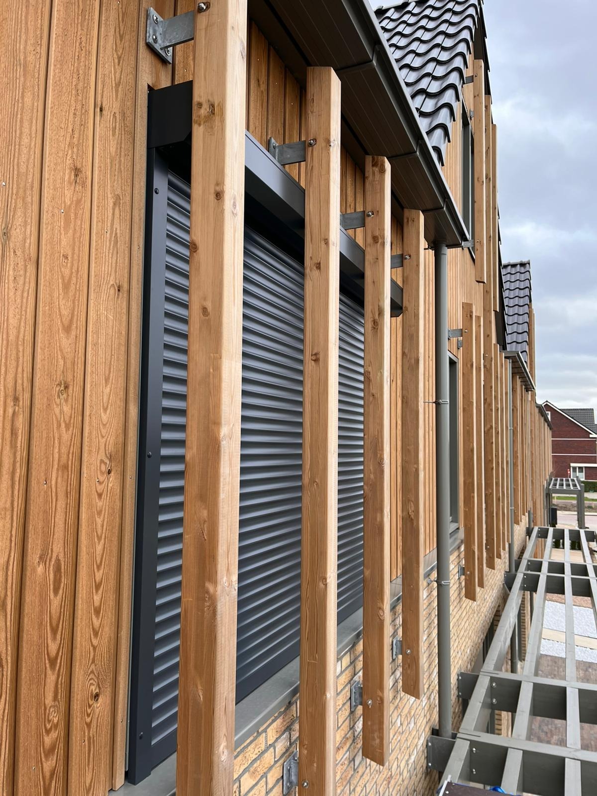 Elewacja budynku z drewnianą okładziną i zamontowaną, zamkniętą roletą zewnętrzną w kolorze grafitowym, widoczne drewniane elementy dekoracyjne.