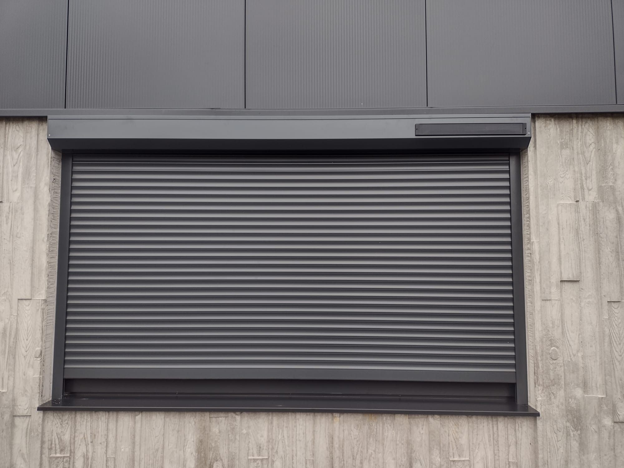 Zasłonięta, szara roleta zewnętrzna na tle betonowej elewacji z drewnianą fakturą. Widoczny fragment metalowej fasady budynku.