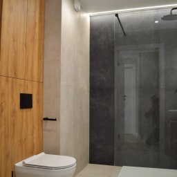 Art Design Grzegorz Kędzierski - Nowoczesna łazienka z drewnianą zabudową, podwieszaną toaletą i kabiną prysznicową ze szklaną ścianką. Minimalistyczny design, beżowe płytki i oświetlenie LED.