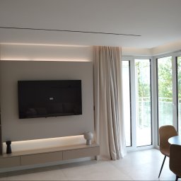 Art Design Grzegorz Kędzierski - Nowoczesny salon z telewizorem na ścianie, beżowymi meblami i dużymi oknami balkonowymi z zasłonami. Minimalistyczny design, jasne kolory, okrągły stół z krzesłami.