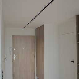 Art Design Grzegorz Kędzierski - Nowoczesny przedpokój z beżową szafą w zabudowie, jasnymi drzwiami wejściowymi i białymi ścianami. Na suficie czarna listwa oświetleniowa.