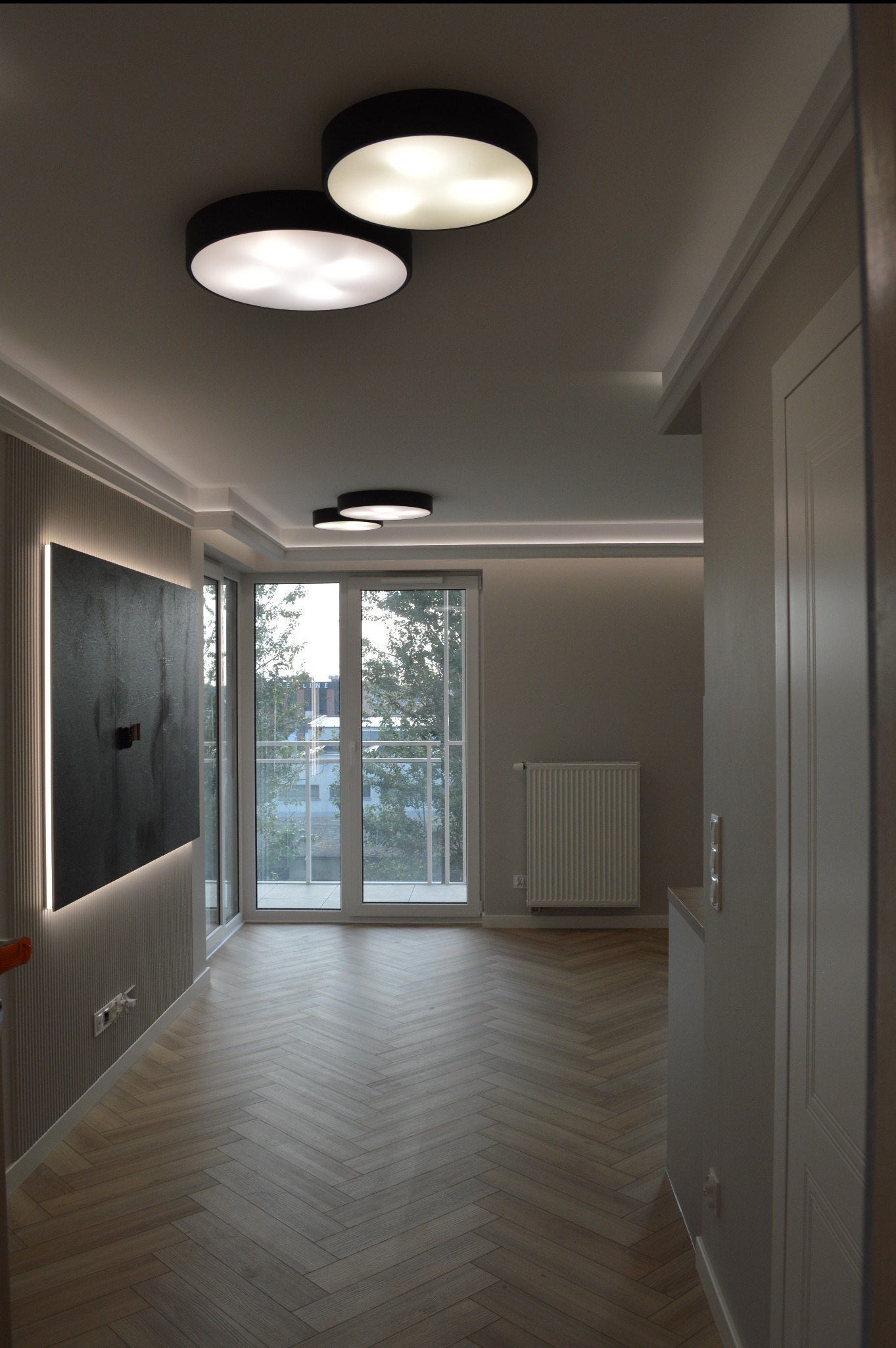 Nowoczesne wnętrze: parkiet w jodełkę, minimalistyczne oświetlenie sufitowe, ściana z dekoracyjną listwą i podświetlany obraz. Jasne, przestronne pomieszczenie.