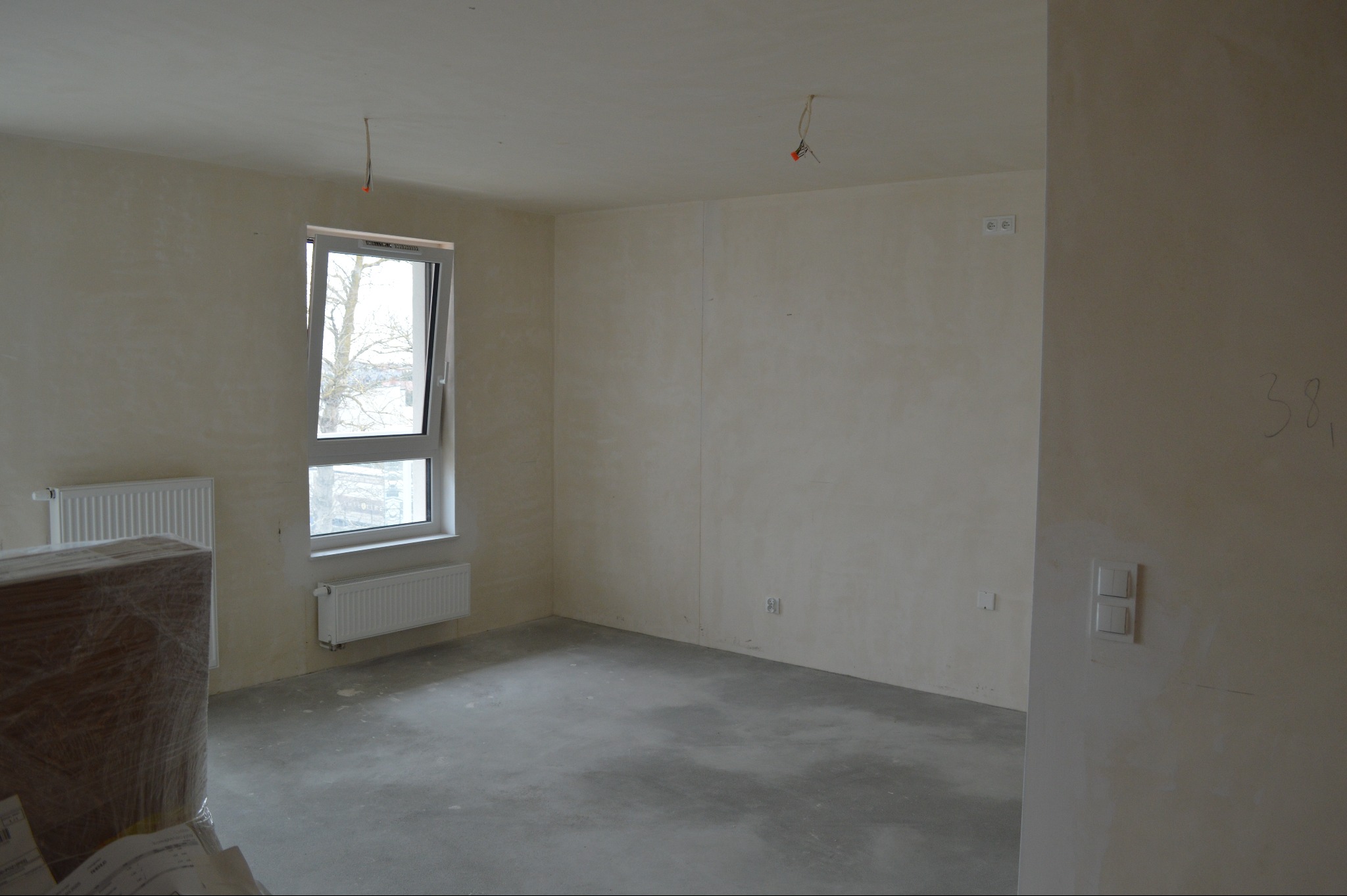 Surowe wnętrze pokoju z oknem i grzejnikami, ściany przygotowane do malowania, widoczne instalacje elektryczne. Podłoga betonowa, w rogu zapakowane elementy.