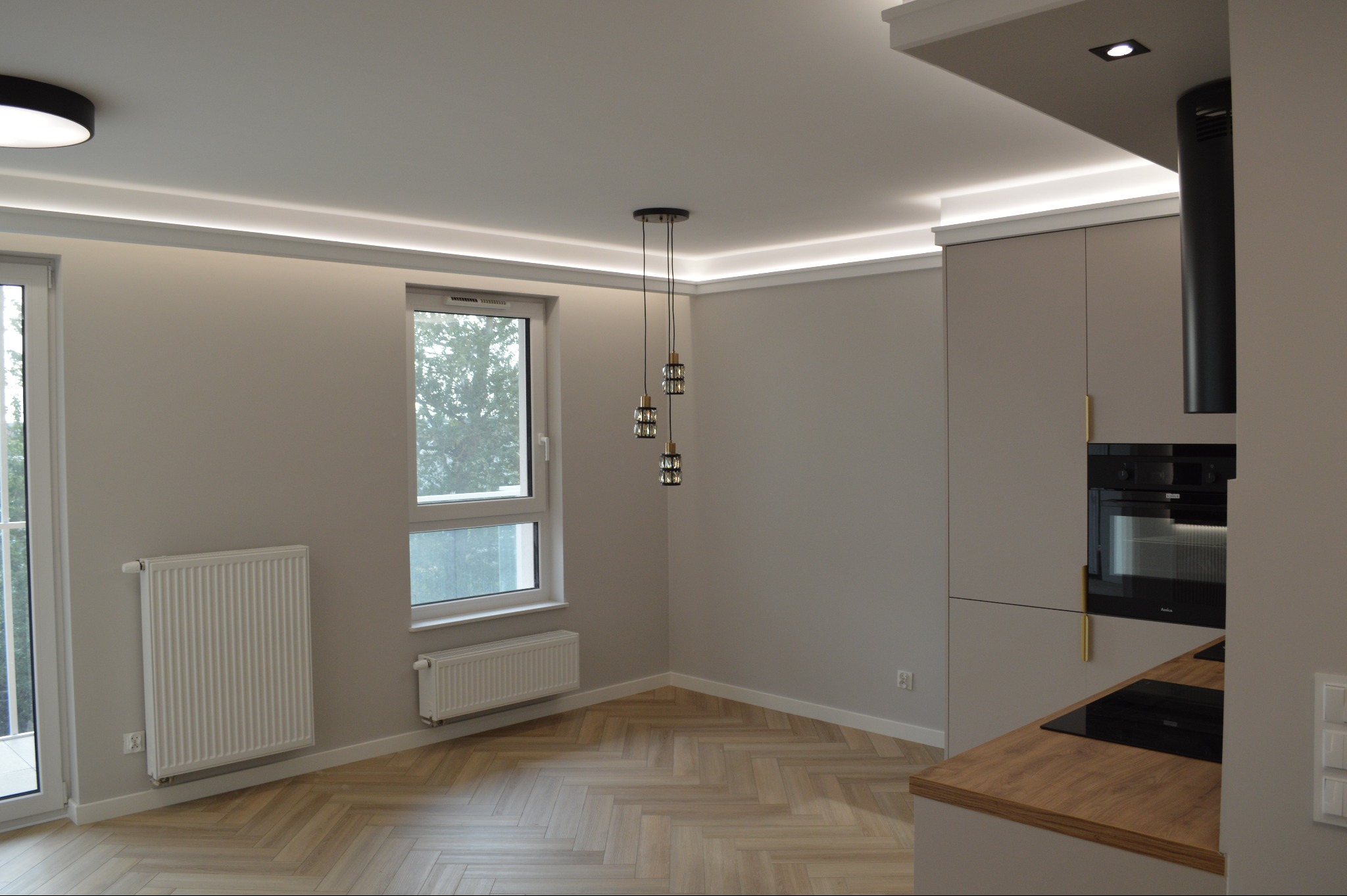 Jasne wnętrze po remoncie: parkiet w jodełkę, szara zabudowa kuchenna z drewnianym blatem, okno z grzejnikiem, nowoczesne oświetlenie sufitowe i wiszące lampy.