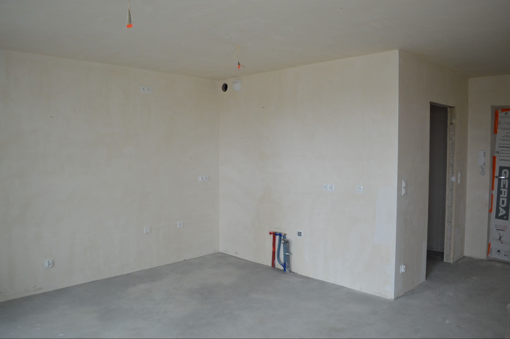 Surowe wnętrze w trakcie wykańczania: gładkie ściany, wylewka, instalacja wodna i elektryczna. Widoczne otwory na gniazdka i rury. Drzwi wejściowe z folią ochronną.