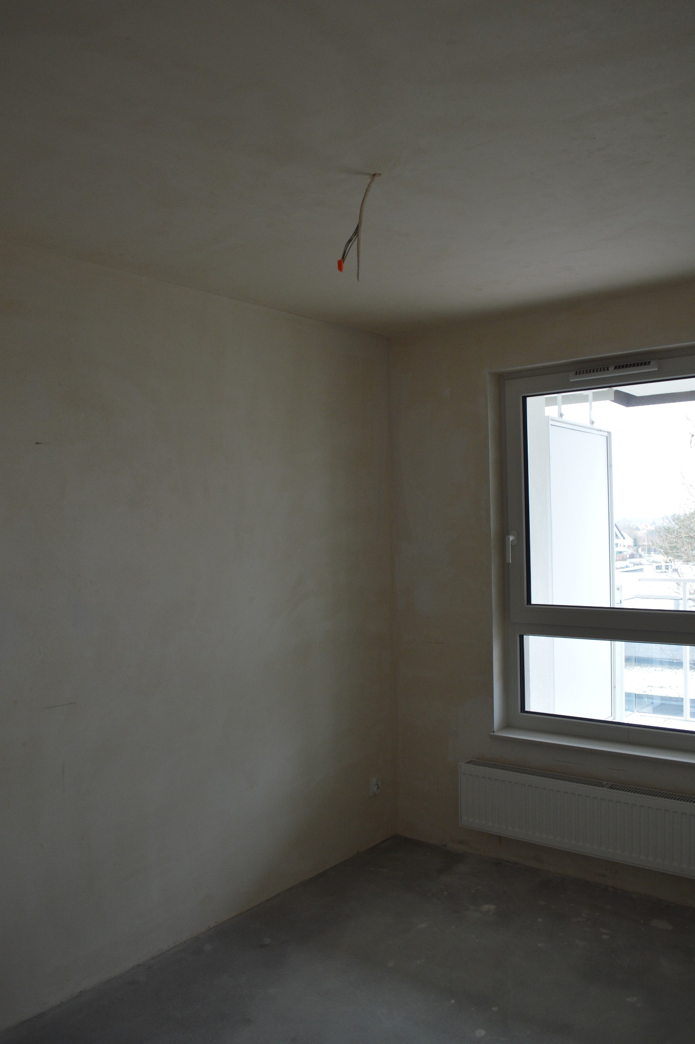 Pusty pokój po gładziowaniu ścian, z oknem i grzejnikiem. Z sufitu zwisają przewody elektryczne. Podłoga betonowa, przygotowana do dalszych prac.