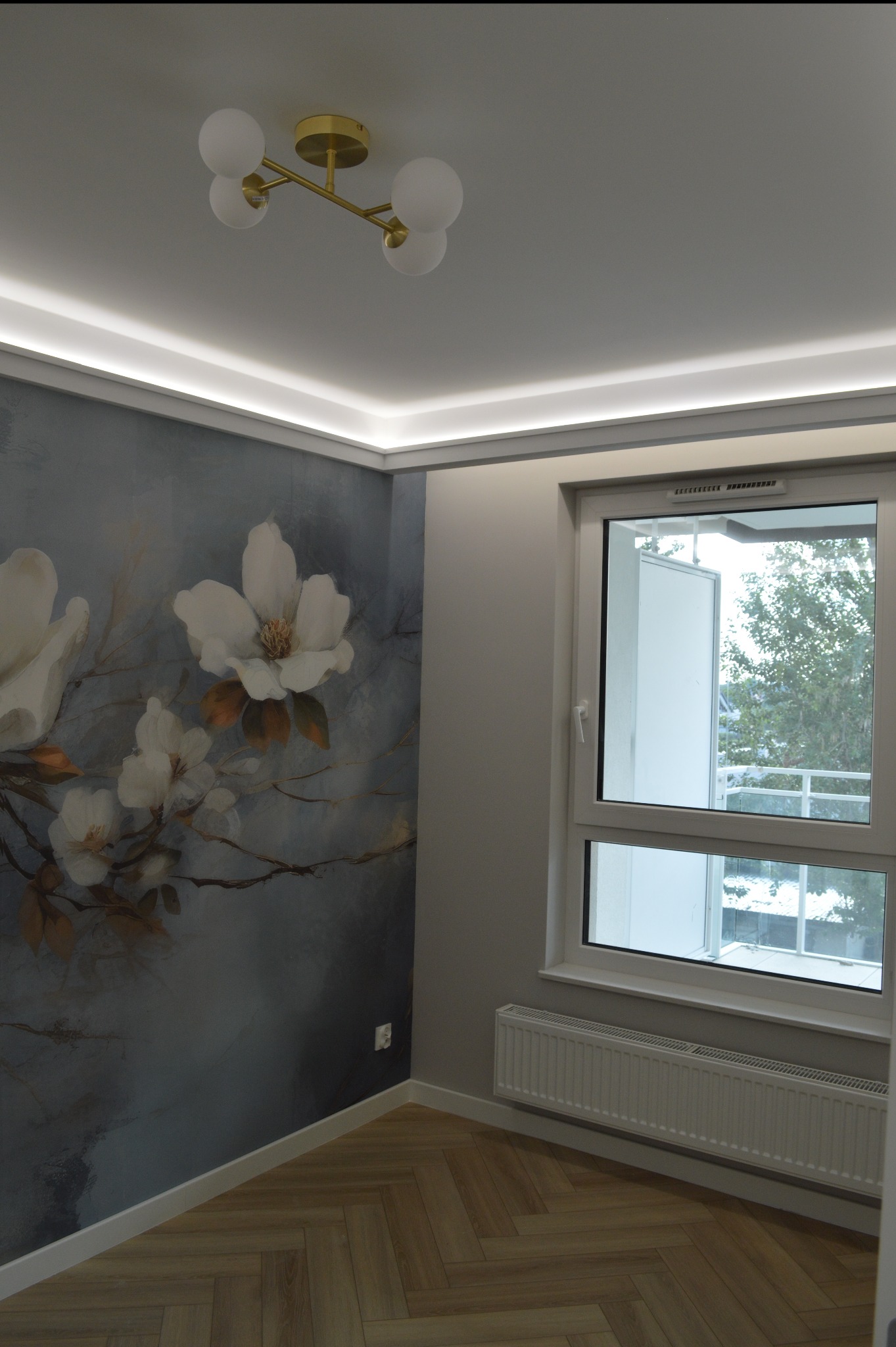 Pokój po remoncie: ściana z motywem kwiatowym, parkiet w jodełkę, okno z widokiem na zieleń, nowoczesna lampa sufitowa i listwy LED pod sufitem.