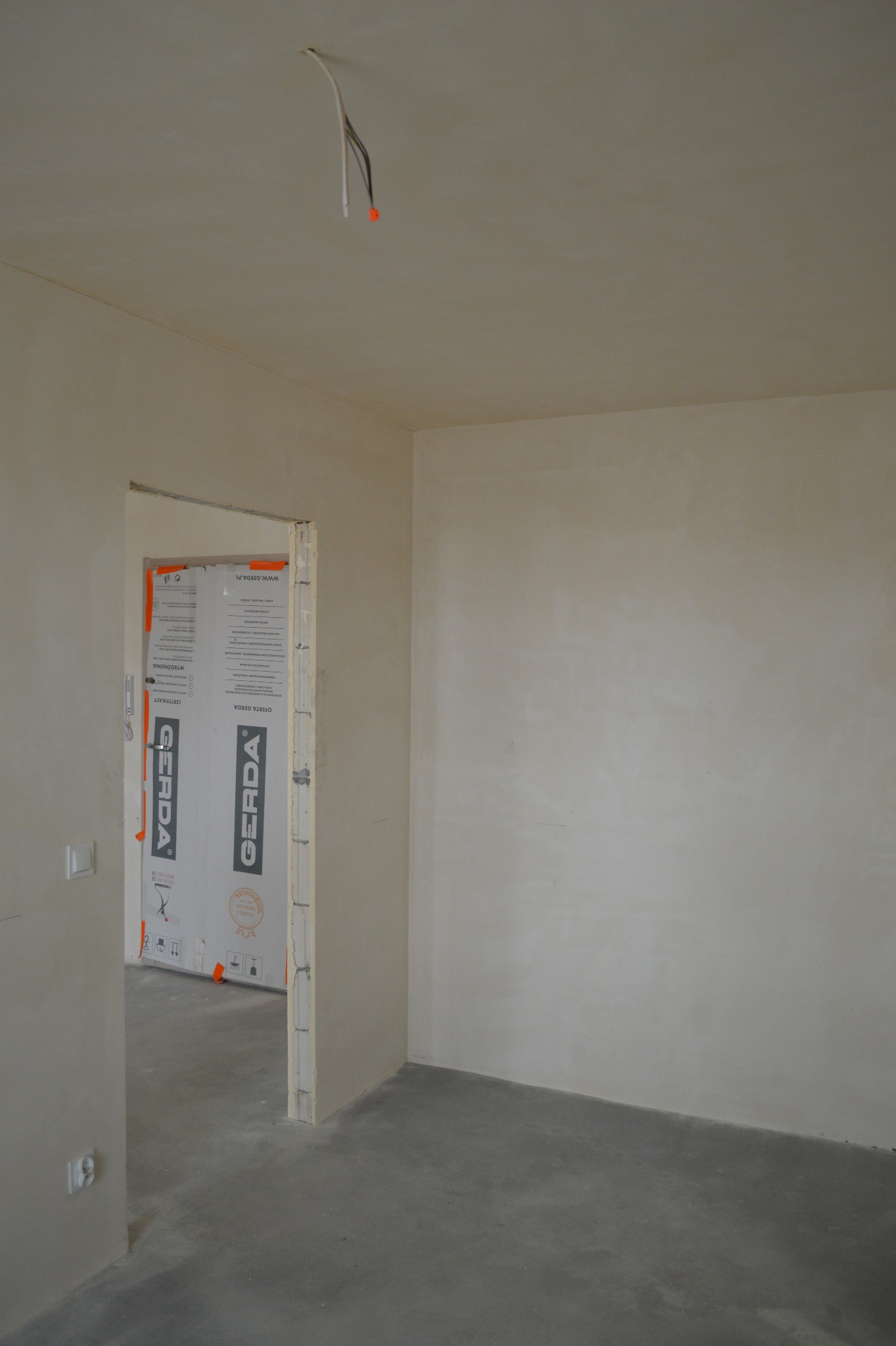 Surowe wnętrze: gładkie, jasne ściany, betonowa podłoga, zabezpieczone futryny drzwiowe. Widoczne kable wystające z sufitu. Minimalistyczny, przygotowany do dalszych prac.