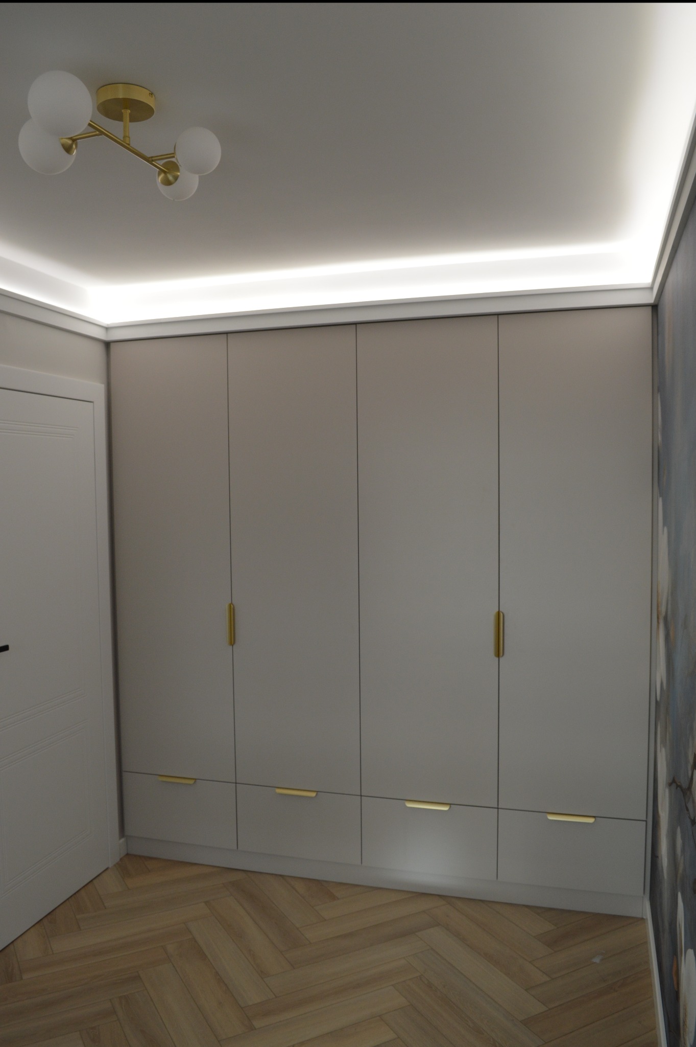 Szara szafa wnękowa z złotymi uchwytami, oświetlenie LED podsufitowe, parkiet w jodełkę. Minimalistyczny design, nowoczesne wykończenie wnętrza.