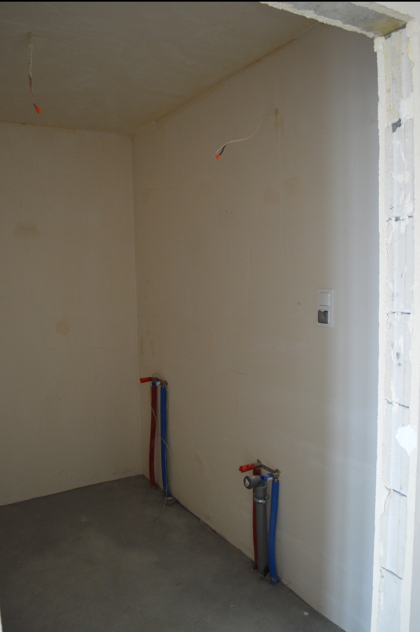 Surowa łazienka w trakcie prac: widoczne instalacje wodne (czerwone i niebieskie rury) przy ścianach, surowy beton na podłodze i wystające kable elektryczne.