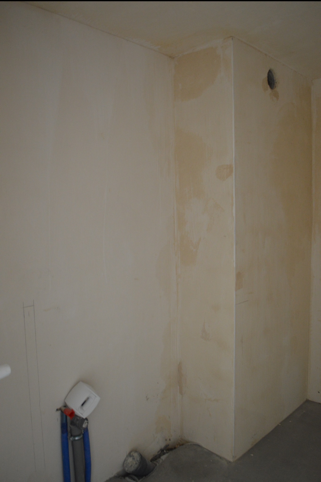 Ściana w trakcie przygotowania do malowania z widocznymi śladami po szpachlowaniu i instalacją hydrauliczną u dołu. Widoczny otwór wentylacyjny w rogu.