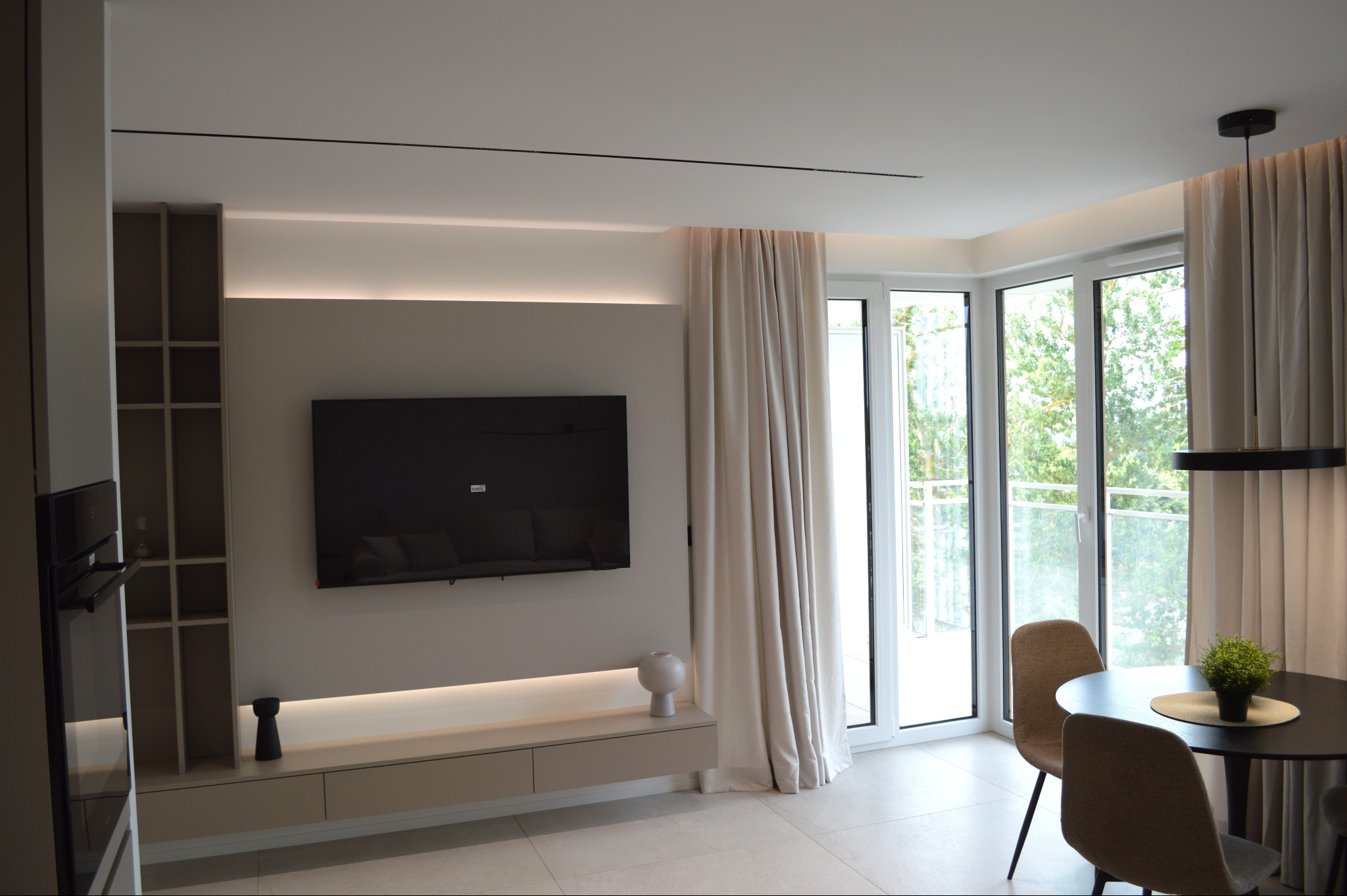 Nowoczesny salon z telewizorem na ścianie, beżowymi meblami i dużymi oknami balkonowymi z zasłonami. Minimalistyczny design, jasne kolory, okrągły stół z krzesłami.