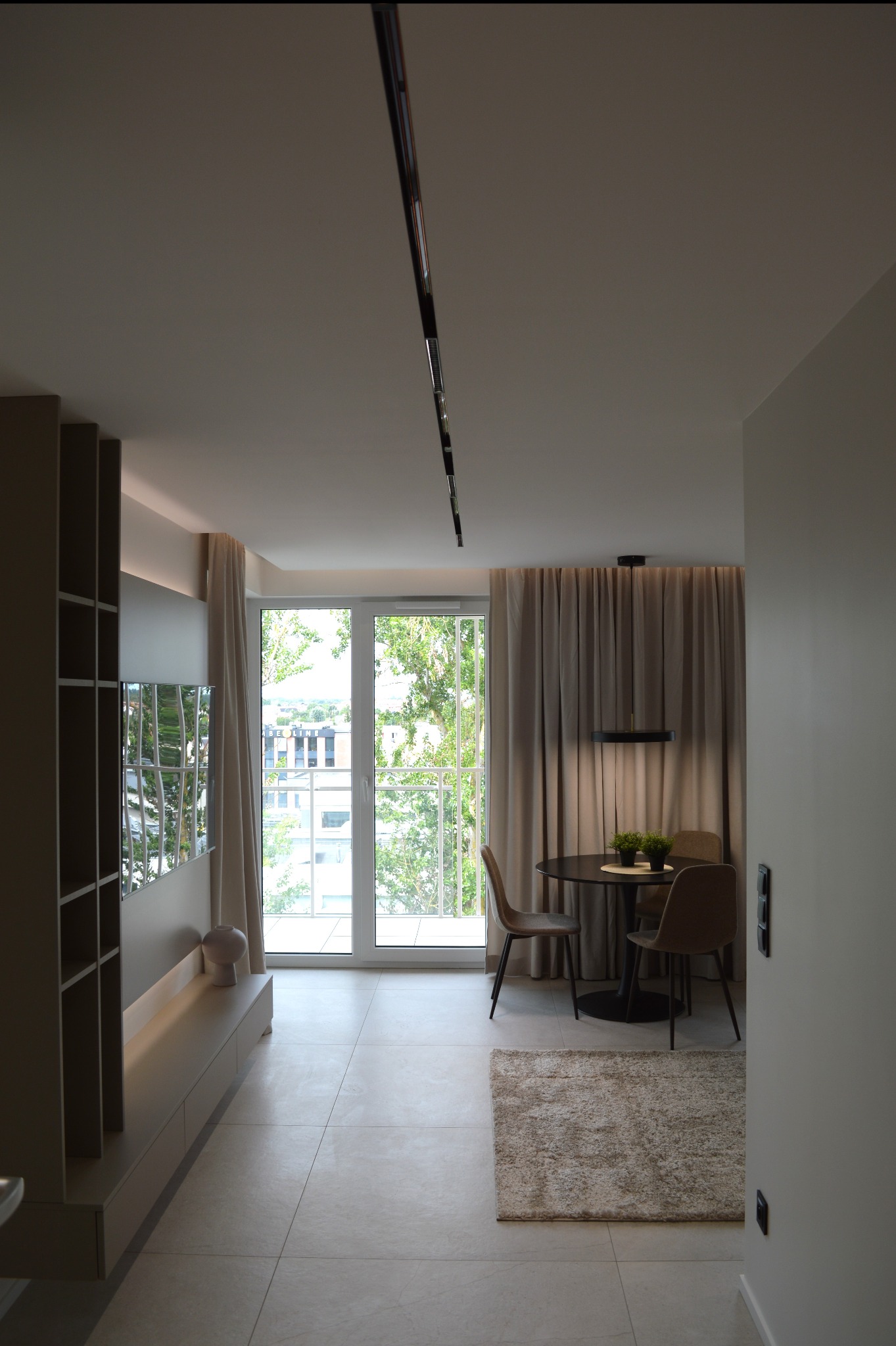 Jasne wnętrze mieszkania z minimalistycznym stołem, krzesłami i dywanem. Duże okno balkonowe z widokiem na zieleń. Nowoczesne oświetlenie sufitowe.