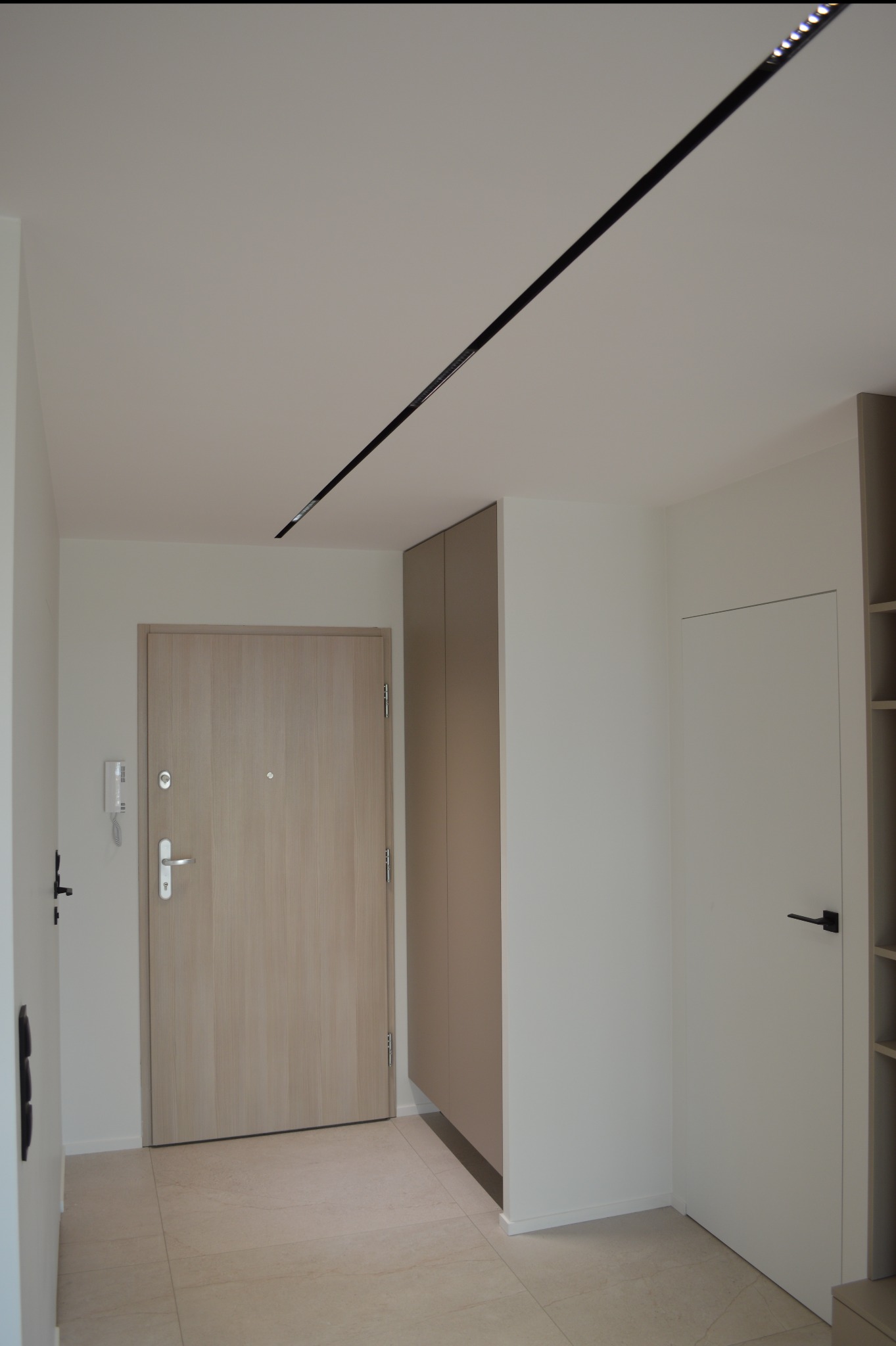 Nowoczesny przedpokój z beżową szafą w zabudowie, jasnymi drzwiami wejściowymi i białymi ścianami. Na suficie czarna listwa oświetleniowa.